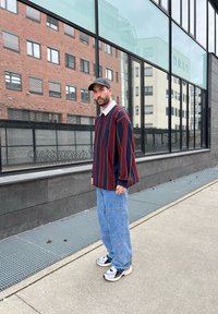 Camicia a maniche lunghe a righe oversized blu navy e bordeaux, jeans baggy azzurri, cappellino nero e sneakers con motivo, in piedi all'esterno accanto a un edificio.