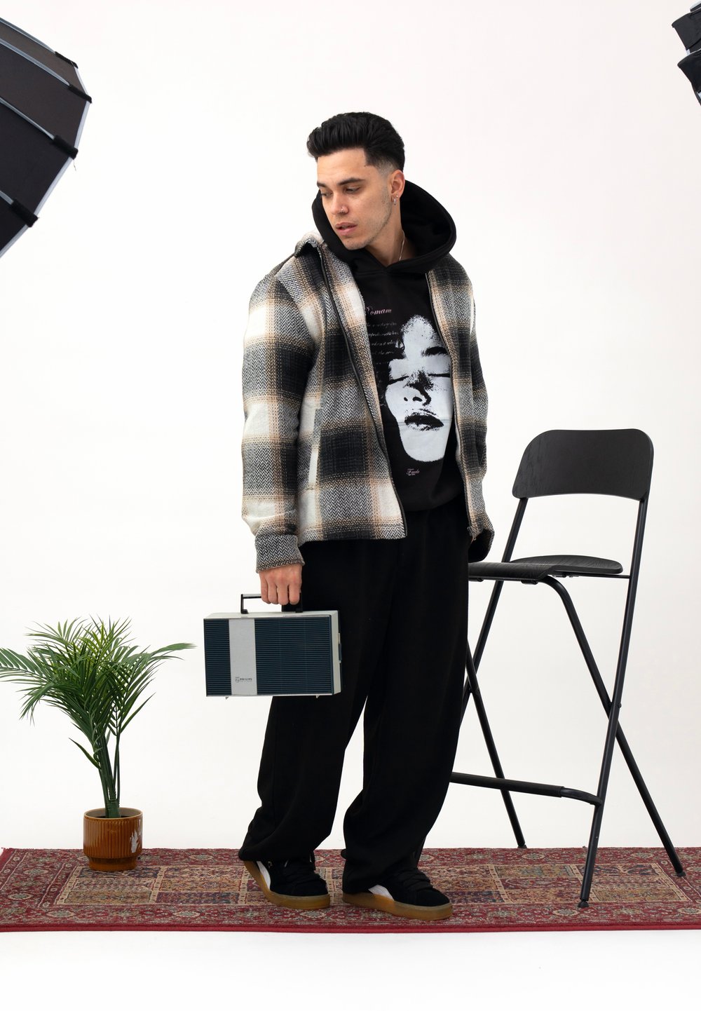 Veste à motif à carreaux gris et crème, sweat à capuche graphique noir, pantalon large noir, tenant une enceinte portable grise, avec une chaise noire à proximité.