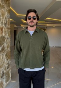 Pulover verde cu model rib pentru o stare confortabilă, cu guler, trei butoane și o croială lejeră, purtat peste o cămașă albă �și asortat cu pantaloni închiși la culoare.