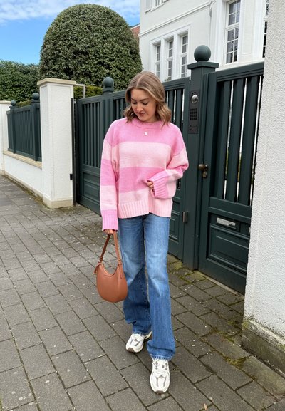 Suéter a rayas rosa, jeans azul claro, bolso beige y zapatillas blancas. Entorno al aire libre con una pasarela pavimentada y una puerta verde al fondo.