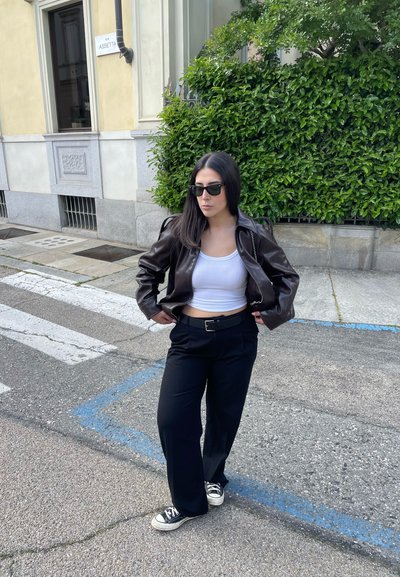Chaqueta de cuero marrón, top corto blanco, pantalones negros con cinturón y zapatillas negras. De pie en una calle con vegetación de fondo.