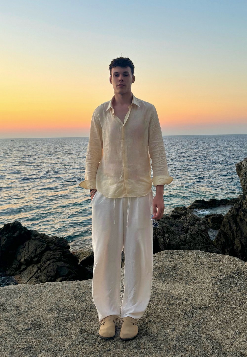 Ανοιχτό κίτρινο λινό πουκάμισο με μακριά μανίκια, λευκές λινές παντελόνες και μπεζ παπούτσια, στέκεται σε έναν βράχο δίπλα στη θάλασσα κατά την διάρκεια του ηλιοβασιλέματος.