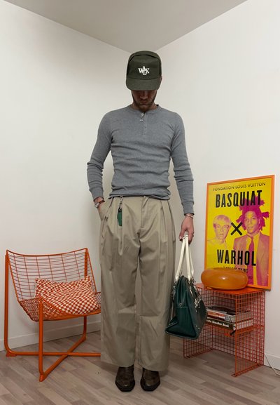 Hombre con camisa gris de manga larga, pantalones anchos beige, gorra verde, sosteniendo una bolsa verde y blanca, de pie cerca de una silla naranja y un póster enmarcado de Basquiat Warhol.