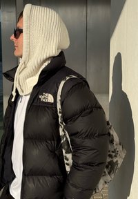 Homme portant une doudoune noire, capuche en maille blanche, lunettes de soleil, et tenant un sac fourre-tout à pois, debout devant un mur clair avec une ombre visible.