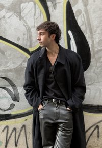 Homme aux cheveux bouclés portant un manteau noir, un pantalon en cuir noir et des lunettes de soleil, se tient les mains dans les poches devant un mur tagué.