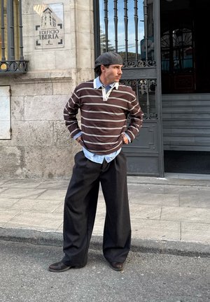 Hombre con boina gris, camisa marrón a rayas, camisa azul con cuello y pantalones anchos negros, de pie con las manos en las caderas frente al Edificio Iberia.