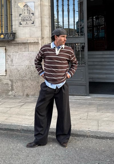 Hombre con boina gris, camisa marrón a rayas, camisa azul con cuello y pantalones anchos negros, de pie con las manos en las caderas frente al Edificio Iberia.