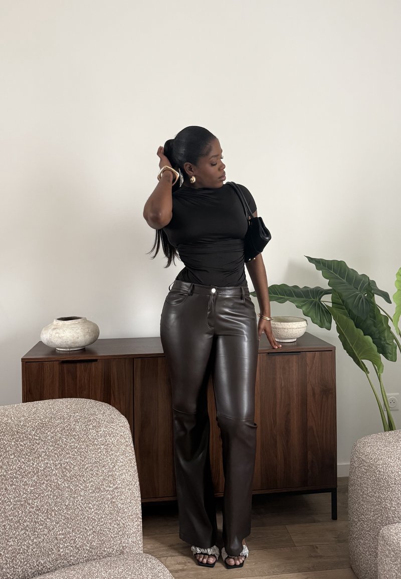 Femme portant un haut noir et un pantalon en cuir sombre se tient dans une pièce moderne, touchant ses cheveux d'une main et reposant l'autre sur une armoire en bois.