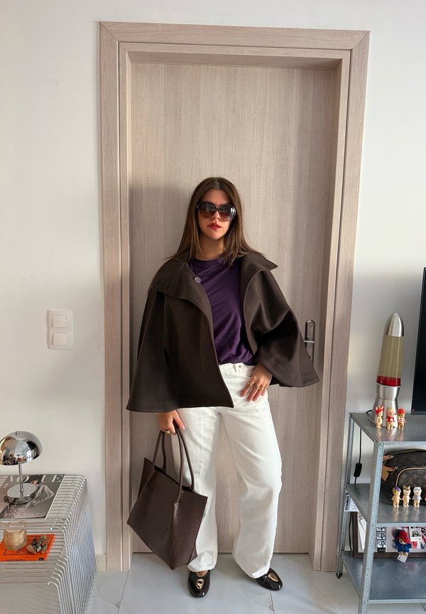 Una mujer con gafas de sol, un abrigo oscuro tipo capa, una prenda superior morada y pantalones blancos posa en una puerta mientras sostiene un bolso oscuro.