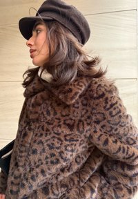 Manteau en fausse fourrure imprimé léopard avec un col, présentant des nuances de brun et de noir. Complété par un chapeau brun foncé et un haut blanc à col haut.