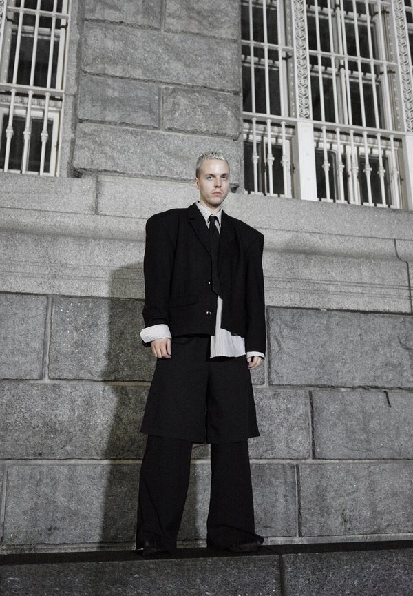 Personne aux cheveux courts et clairs portant une veste de costume noire surdimensionnée et un pantalon large, debout contre un mur en pierre avec de grandes fenêtres à barreaux.
