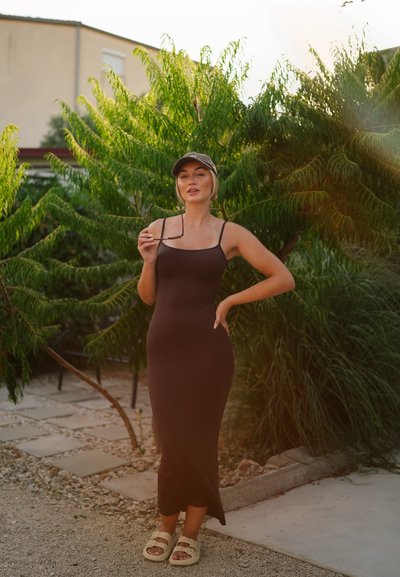 Vestido marrón ajustado con tirantes finos, que llega a la mitad de la pantorrilla. Acompañado de una gorra y sandalias ligeras, sobre un fondo de vegetación exuberante y un camino de piedras.