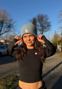 Jeune femme ajustant un bonnet gris LA, portant un haut noir long à manches longues raccourci, debout sur un trottoir ensoleillé avec des arbres et des voitures flous en arrière-plan.