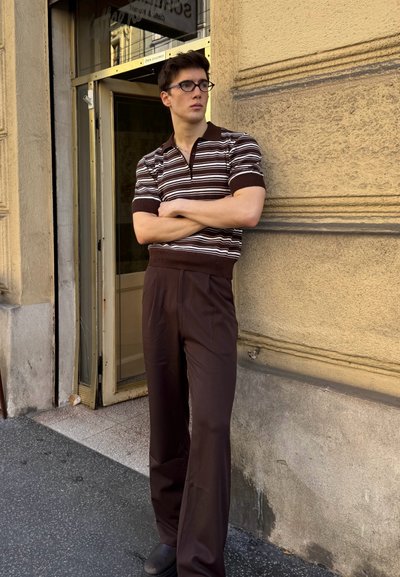 Camisa polo de rayas marrones con mangas cortas, combinada con pantalones de pierna ancha marrones, de pie contra una pared texturizada cerca de una puerta.