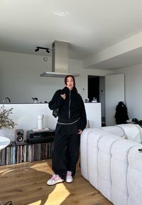 Μαύρο φούτερ με φερμουάρ πάνω από μαύρη στρωμένη μπλούζα, συνδυασμένο με φαρδιά μαύρα παντελόνια και λευκά αθλητικά παπούτσια με ροζ λεπτομέρειες. Μινιμαλιστική κουζίνα στο παρασκήνιο.