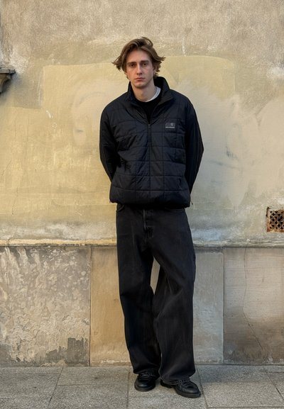 Joven con cabello castaño claro está apoyado contra una pared beige texturizada, lleva una chaqueta negra acolchada, pantalones negros holgados y zapatos negros.