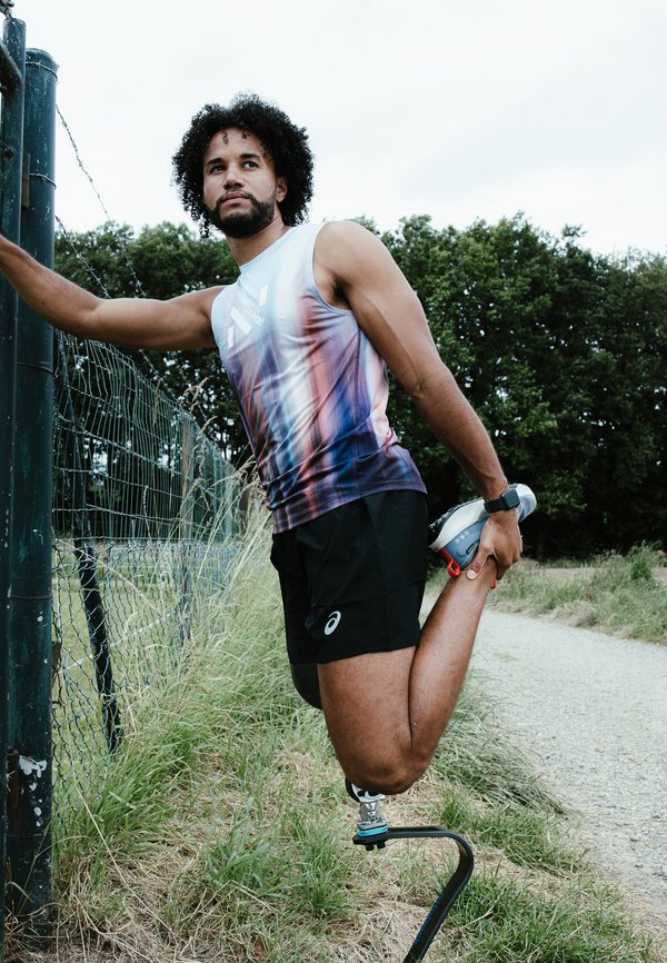 Un atleta estirándose, vestido con una camiseta sin mangas de color púrpura y blanco con un patrón degradado y pantalones cortos negros, con una pierna prostética y un zapato en la mano.