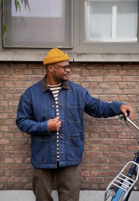 Denim jack met een bruine kraag, bruine corduroy broek en een mosterdkleurige beanie. Onder de jack is een gestreept shirt te zien, terwijl hij naast een fiets staat.