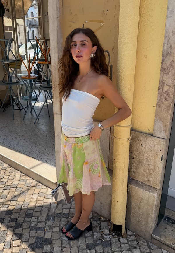 Mujer con el pelo largo, vestida con un top blanco sin tirantes, falda verde y rosa, sandalias negras y pendientes plateados, de pie junto a una pared amarilla.