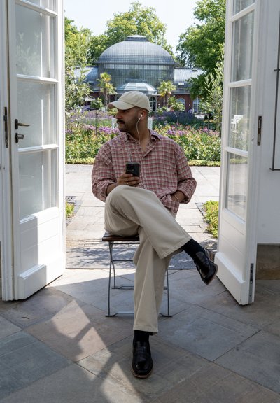Un hombre está sentado en una silla, vistiendo una camisa de cuadros, pantalones beige y zapatos negros, sosteniendo un teléfono con auriculares. Puertas abiertas enmarcan un jardín.