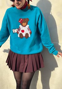 Sweat-shirt turquoise avec un écusson d'ours en robe blanche, accents à carreaux et un cœur. Associé à une jupe plissée marron.