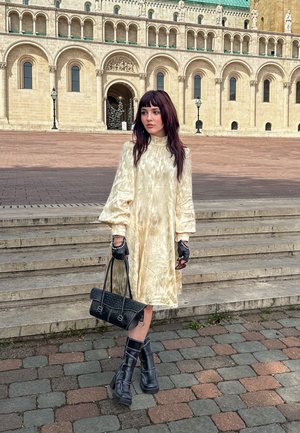 D długa, bladożółta satynowa sukienka z bufiastymi rękawami i wysokim kołnierzem. Czarnie rękawiczki, czarne botki na platformie sięgające kostki oraz fakturowana czarna torebka.