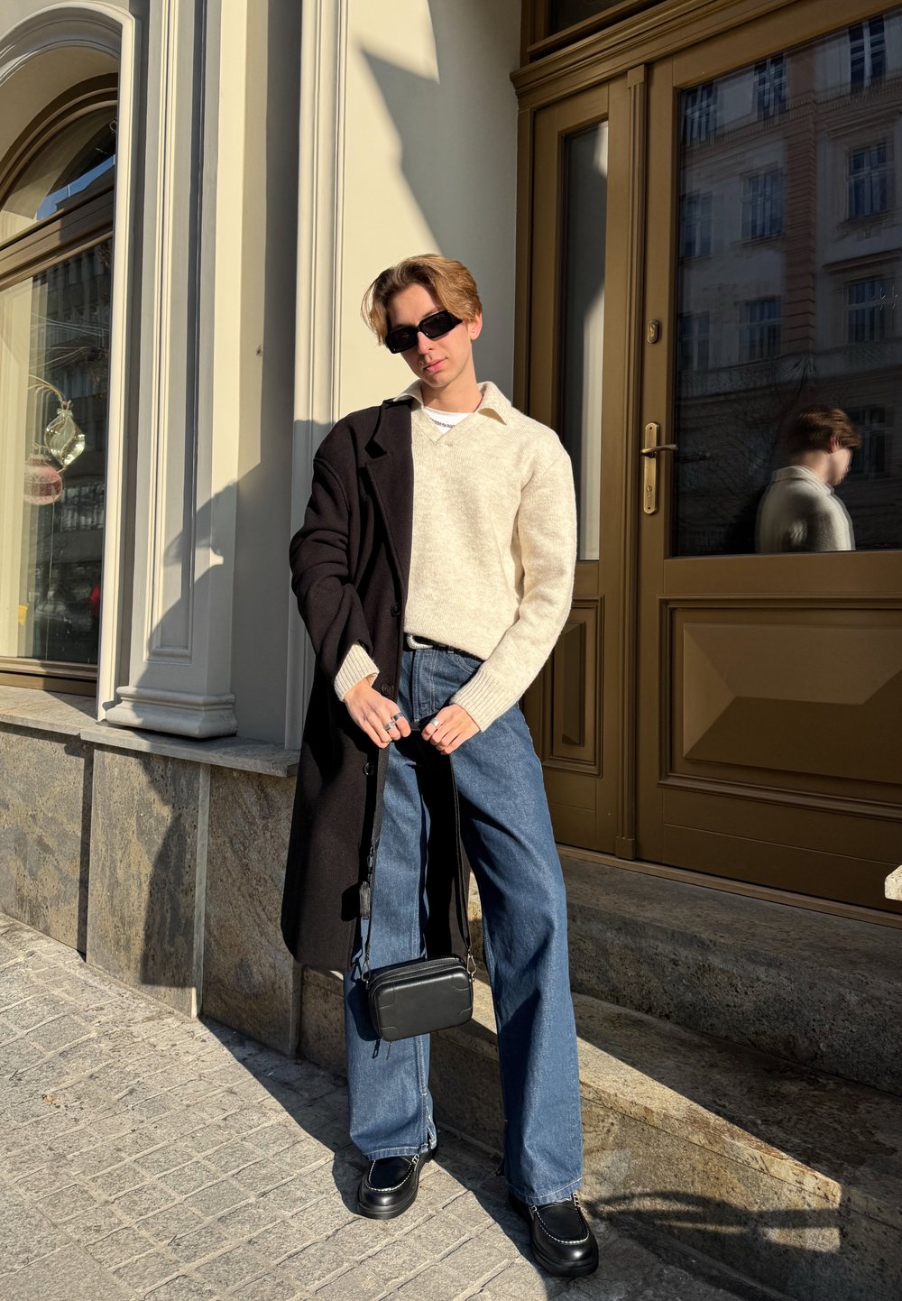 Καθημερινό ντύσιμο που περιλαμβάνει ένα κρεμ πουλόβερ, μαύρο μακρύ παλτό, φαρδύ τζιν σε μπλε απόχρωση και μαύρη τσάντα χιαστί, με τετράγωνα γυαλιά ηλίου και ογκώδη μαύρα παπούτσια.