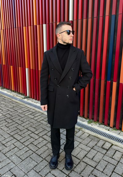Abrigo negro de doble botonadura con grandes botones, llevado sobre un cuello de tortuga negro. Combinado con pantalones negros ajustados y zapatos negros resistentes. El fondo presenta rayas verticales de colores.
