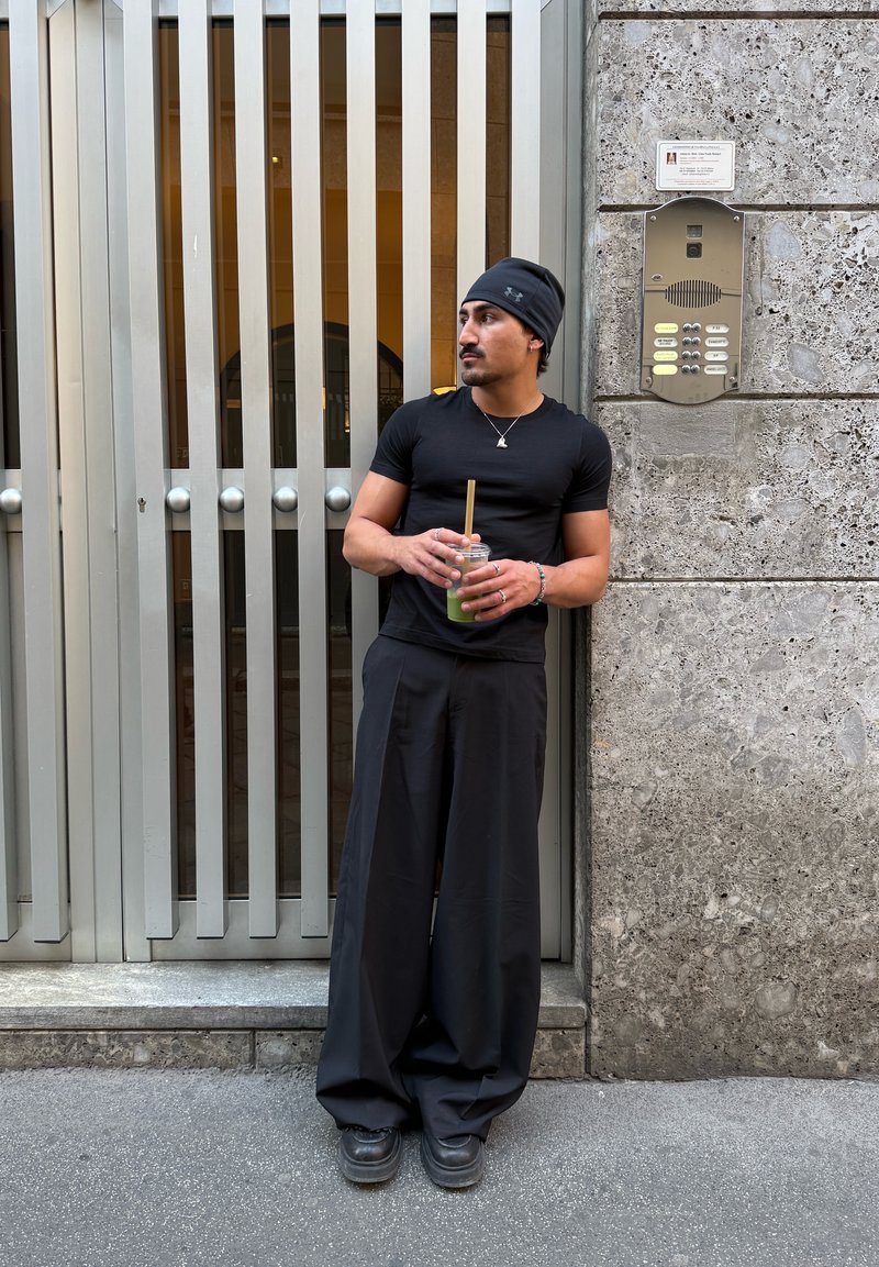 T-shirt preta ajustada, calças largas pretas, gorro preto. A segurar um copo transparente com um canudo. De pé contra uma parede texturizada cinza e um portão.