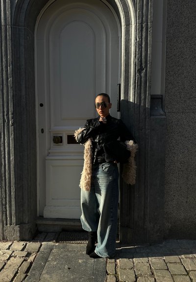 Persona con gafas de sol, chaqueta negra y vaqueros anchos, de pie junto a una puerta arqueada con marco de piedra, sombreada por la luz del sol.