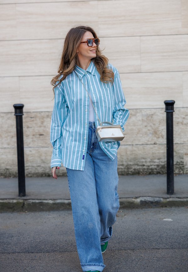 Femeie purtând o cămașă în dungi albastre și albe, blugi cu picioare largi, ochelari de soare și o geantă mică albă pe umăr, mergând pe o stradă asfaltată.