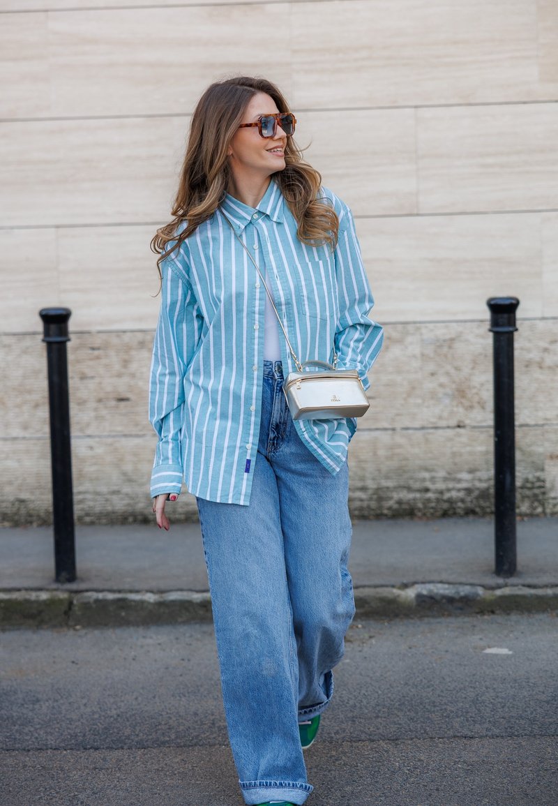Femme portant une chemise à rayures bleues et blanches, un jean à jambes larges, des lunettes de soleil et un petit sac blanc porté en bandoulière, marchant dans une rue pavée.