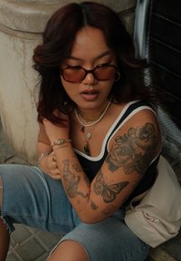 Óculos de sol castanhos, uma blusa sem mangas azul-marinho com bordas brancas, calções de ganga e várias tatuagens. Colares com diversos materiais e pendentes. Bolsa leve.
