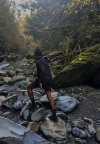 Atletische man in donkere kleding die een rotsachtige rivierbedding doorkruist. Belangrijke kenmerken zijn gladde grijze stenen, met mos bedekte rotsen en omringende bomen in herfstkleuren.