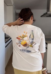 Krém színű póló, amelyen nyomtatott minta látható egy kávéscsészéről, bagetről, citromról, vizespalackról, tulipánokról és dobókockáról.