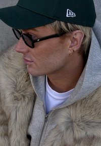 Persoon met een groene pet, zwarte bril en een grijze capuchontrui met rits en een bontkraag, aangevuld met gouden oorbellen.