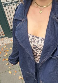 Chaqueta de mezclilla oscura con solapas anchas, dos bolsillos delanteros y costuras naranjas, superpuesta a una blusa de estampado de leopardo claro, sobre un fondo de pavimento.