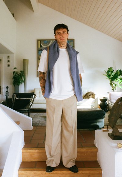 Chaleco de punto azul sobre una camiseta blanca, pantalones de pierna ancha beige y zapatos negros. De pie en unas escaleras de madera en un espacio interior moderno.