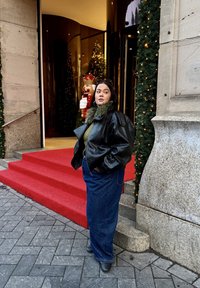 Jachetă bomber din piele neagră, pulover verde din tricot și blugi evazați albaștri. Trepte de covor roșu și decorațiuni festive în fundal.