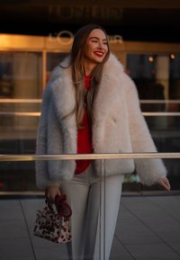 Faux fur krämfärgad jacka med bred krage, röd tröja och vita byxor. Bär på en liten vit väska med körsbärsmönster.