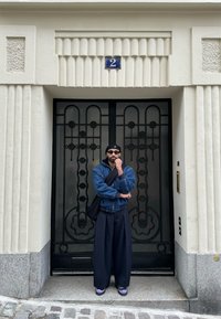 Melnā kaligā metalizētā durvju ar ģeometriskiem akcentiem, augsta balta ēka ar teksturētu virsmu, indivīds tumšā džinsa jakā un platās biksēs.