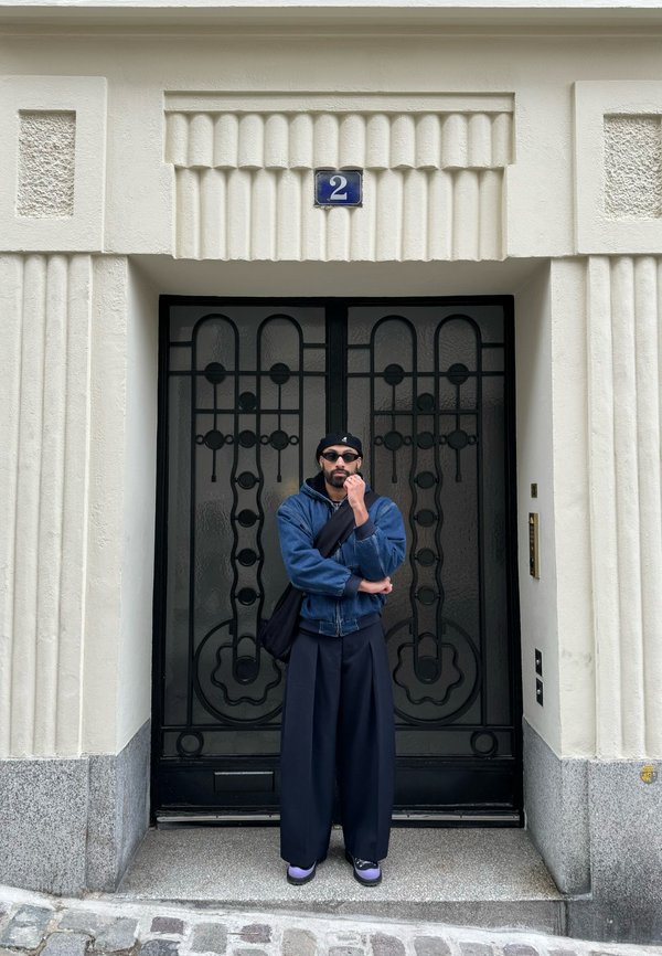 Svart smijernsdør med geometrisk utsmykning, høy hvit bygning med teksturert overflate, individ i mørk denimjakke og vide bukser.