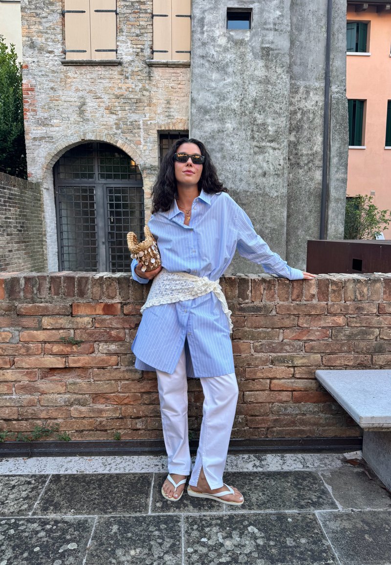 Gestreiftes blaues Oversized-Shirt mit weißen Hosen, einem Spitzen-Gürtel und weißen Flip-Flops. Hält ein strukturiertes Accessoire. Hintergrund aus Ziegelmauer.
