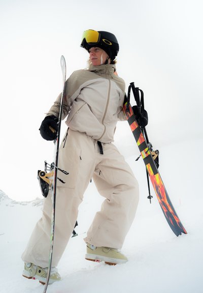 Esquiador con chaqueta y pantalones beige sostiene esquís con diseño de llamas, lleva casco negro y gafas amarillas, de pie en una pendiente nevada.