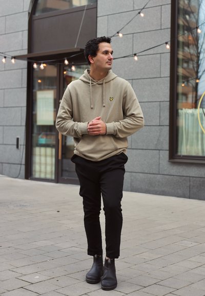 Sudadera beige con cordones y logo amarillo, combinada con pantalones negros y botas negras hasta el tobillo, con un fondo urbano.