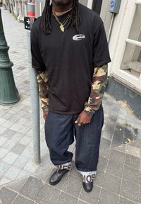 Uomo con dreadlocks che indossa una maglietta nera "Quotrell" sopra maniche mimetiche, jeans larghi scuri con i risvolti, scarpe nere e collane d'oro, in piedi sul marciapiede.