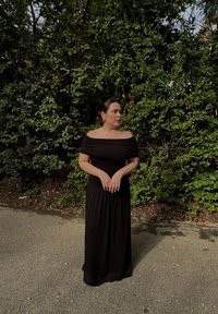 Robe maxi noire à épaules dénudées en tissu fluide, avec un corsage ajusté et une jupe ample. Le modèle se tient devant un feuillage vert.