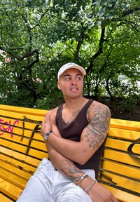 Top de tirantes color canela, pantalones gris claro, una gorra clara y varios tatuajes. Sentado en un banco amarillo con grafitis negros y follaje verde al fondo.