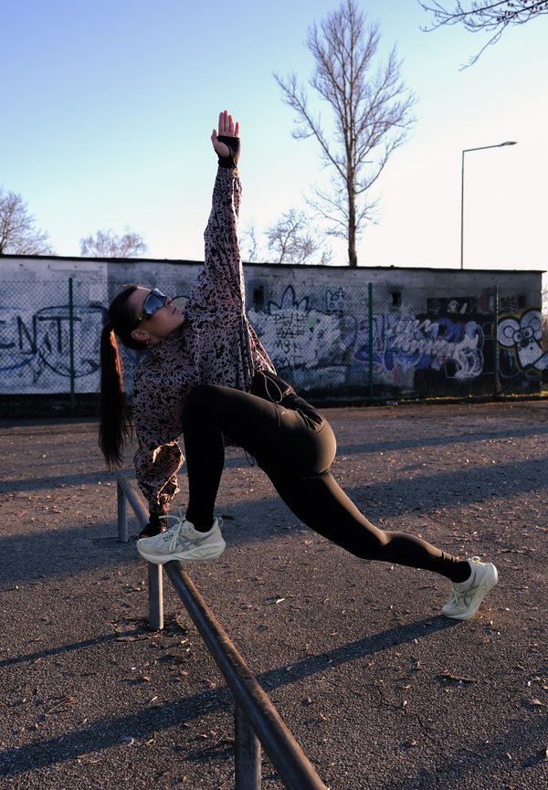 Mujer con ropa deportiva estirándose sobre una barandilla de metal en un entorno urbano al aire libre con una pared con grafitis y árboles sin hojas al atardecer.