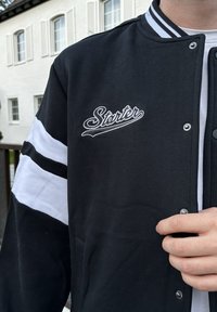 Schwarze Collegejacke mit weißen Streifen an den Ärmeln, "Starter" in Weiß auf der Brust gestickt, Druckknöpfe und eine Baumwollstruktur.